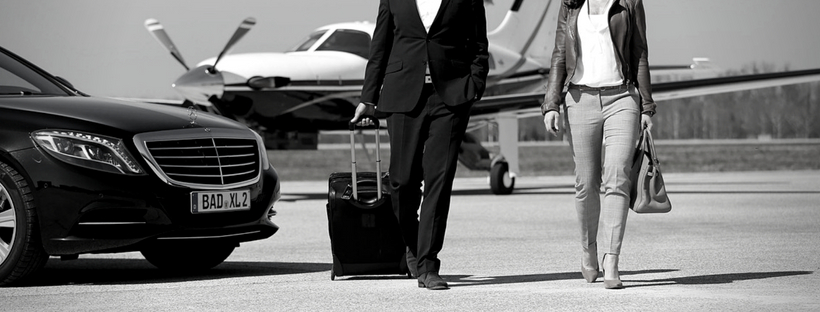 Ein Mann mit einem Rollkoffer und eine Frau mit Handtasche vor einem Flugzeug und einer Mercedes Limousine.